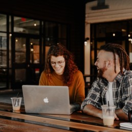 Two young people sitting down at a cafe having a coffee and laughing in front of a computer